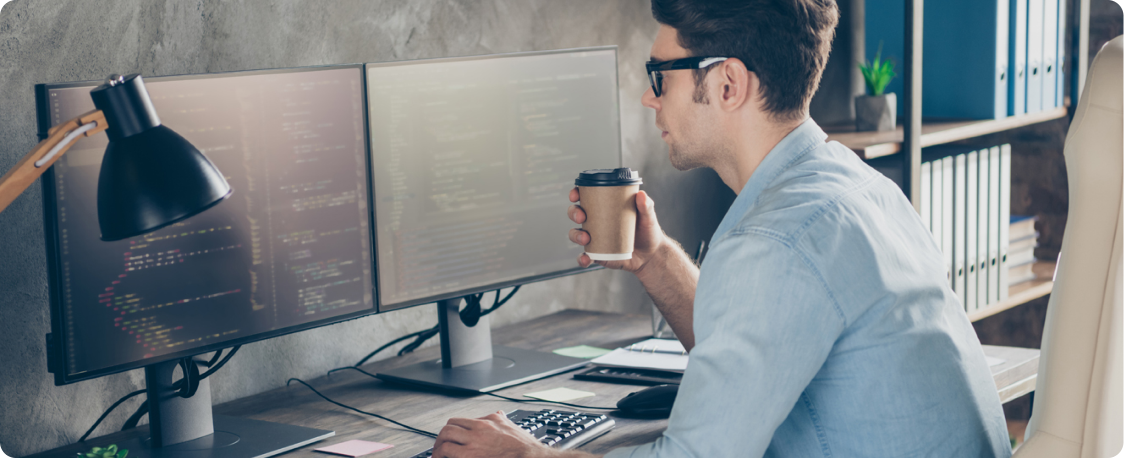 Мужчина в синей рубашке держит кофе в бумажном стаканчике, работая за настольным компьютером.