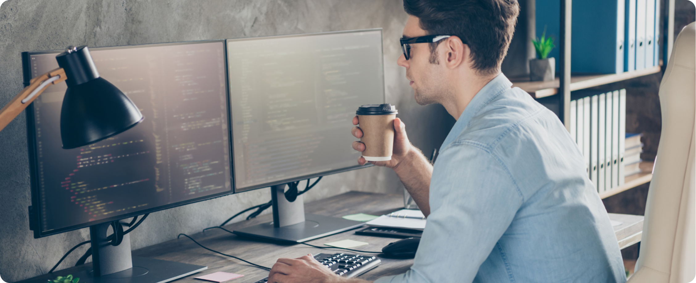 Een man in een blauw overhemd houdt een papieren koffiebeker vast terwijl hij op een desktopcomputer werkt.